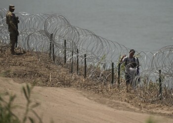 Migrants walks along concertina wire past a guardsman after crossing the Rio Grande from Mexico near the site where large buoys are being deployed to be used as a border barrier in Eagle Pass, Texas, Wednesday, July 12, 2023. The floating barrier is being deployed in an effort to block migrants from entering Texas from Mexico. (AP Photo/Eric Gay)