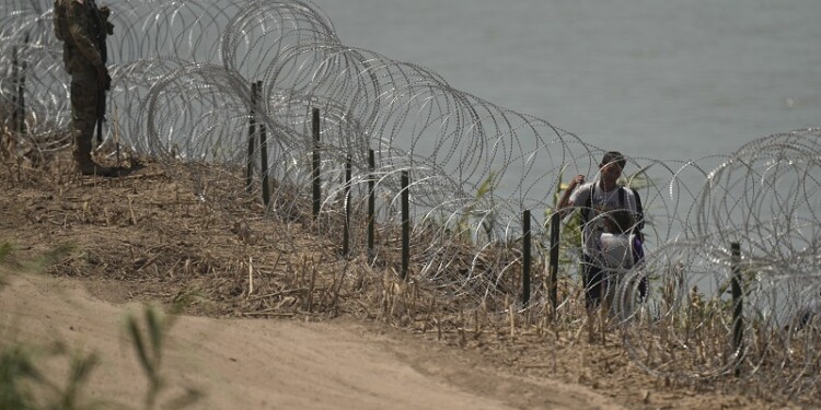 Migrants walks along concertina wire past a guardsman after crossing the Rio Grande from Mexico near the site where large buoys are being deployed to be used as a border barrier in Eagle Pass, Texas, Wednesday, July 12, 2023. The floating barrier is being deployed in an effort to block migrants from entering Texas from Mexico. (AP Photo/Eric Gay)