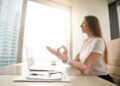 Calm peaceful businesswoman practicing yoga at office desk sitting near laptop, hand in chin mudra yogic gesture, meditation at work, online yoga exercises, practicing breathing, no stress, side view