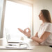 Calm peaceful businesswoman practicing yoga at office desk sitting near laptop, hand in chin mudra yogic gesture, meditation at work, online yoga exercises, practicing breathing, no stress, side view