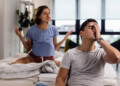 Young man feeling stressed out and having a headache while his girlfriend is arguing with him in the bedroom.